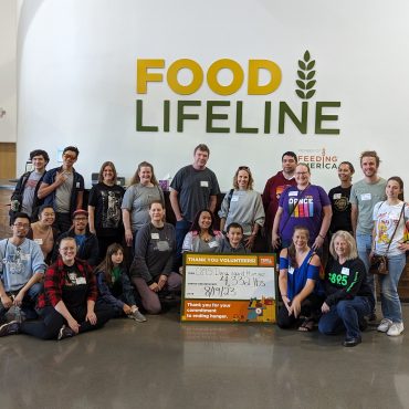 A large group of volunteers standing in front of a sign that says "Food Lifeline" with a smaller sign saying how many tons of food they packed.