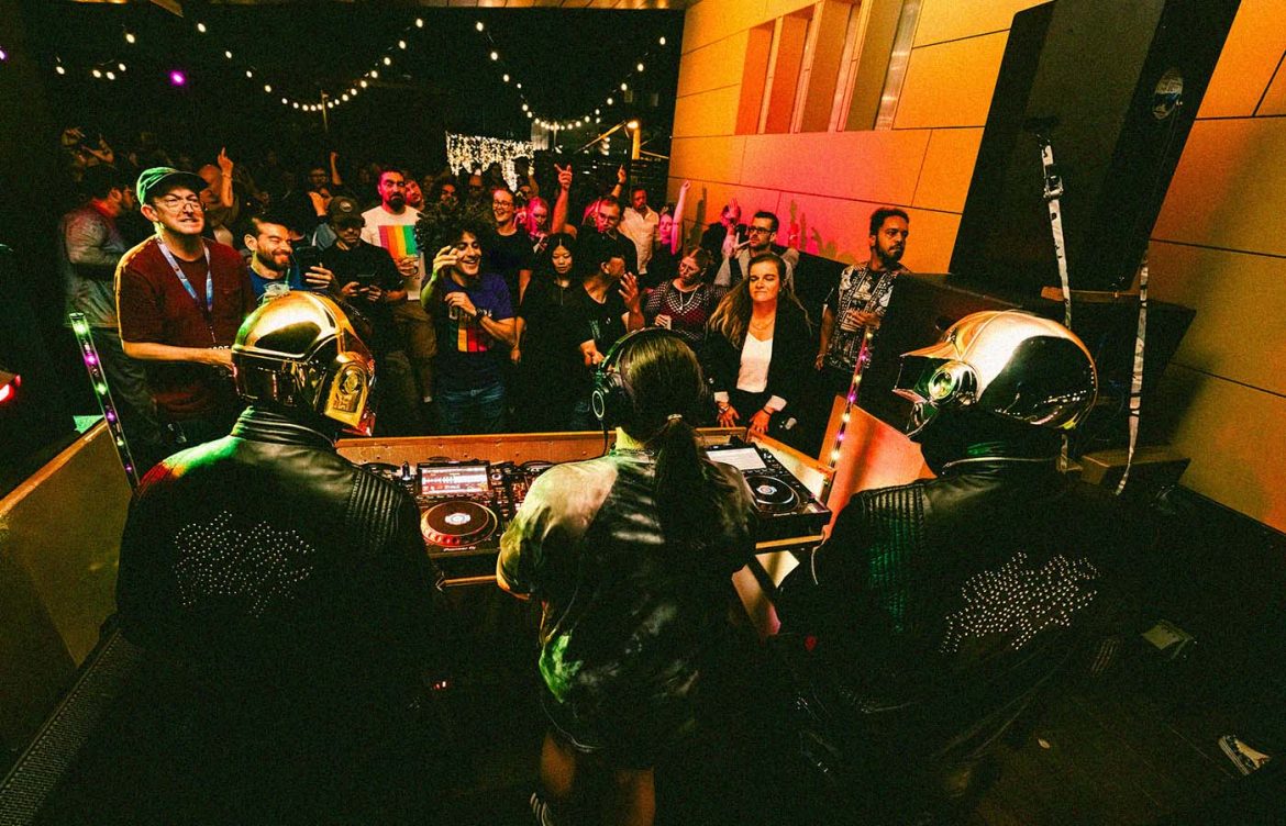 A DJ performance in a dimly lit room. Two individuals wearing gold helmets, reminiscent of Daft Punk, stand at the DJ booth. They are performing to a lively crowd, with people visible in the background enjoying the music. The room is decorated with string lights, adding to the festive atmosphere.