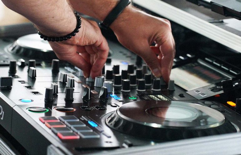 A person adjusting controls on a professional music mixing console, focusing on various knobs and sliders. Their wrist displays a smartwatch, and a tattoo is visible on their arm. The equipment is detailed and technical, suggesting a DJ setup.