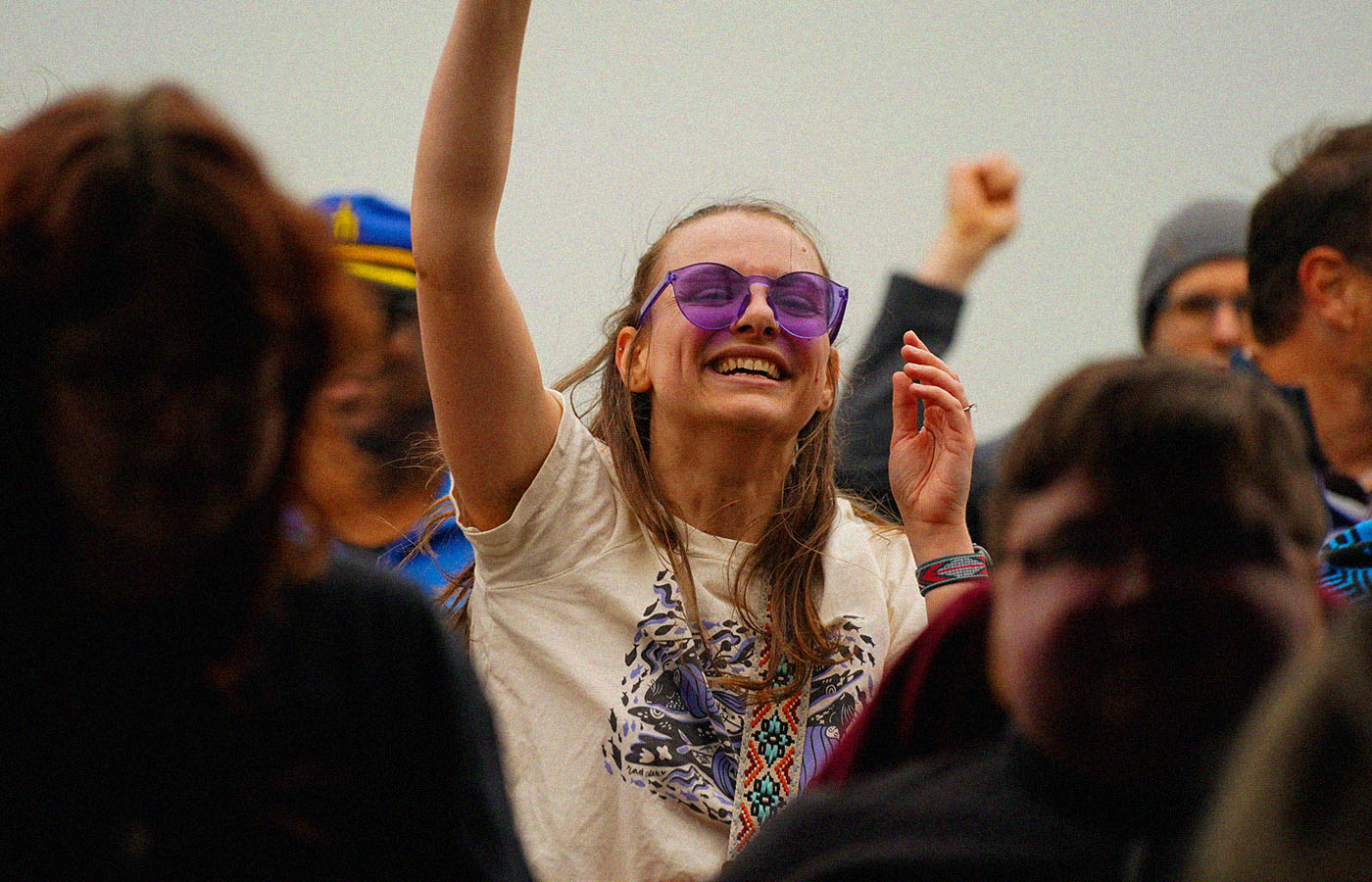 A person wearing purple sunglasses and a graphic t-shirt is joyfully raising an arm amidst a crowd. The background includes other people, some of whom are also visibly excited.