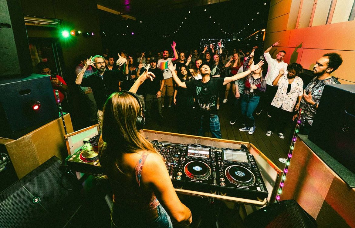 A DJ is performing in front of a lively crowd under colorful lights in a club-like setting. The audience is engaged and raising their hands, with a backdrop of string lights and vibrant decor.