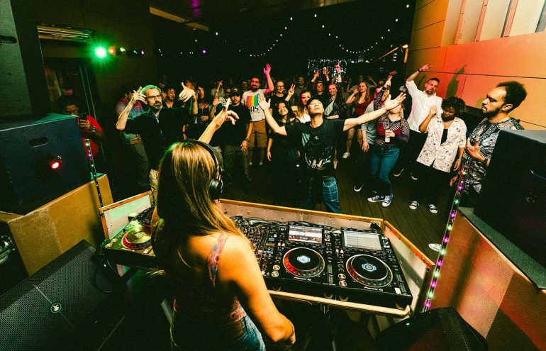 A DJ is performing in front of a lively crowd under colorful lights in a club-like setting. The audience is engaged and raising their hands, with a backdrop of string lights and vibrant decor.