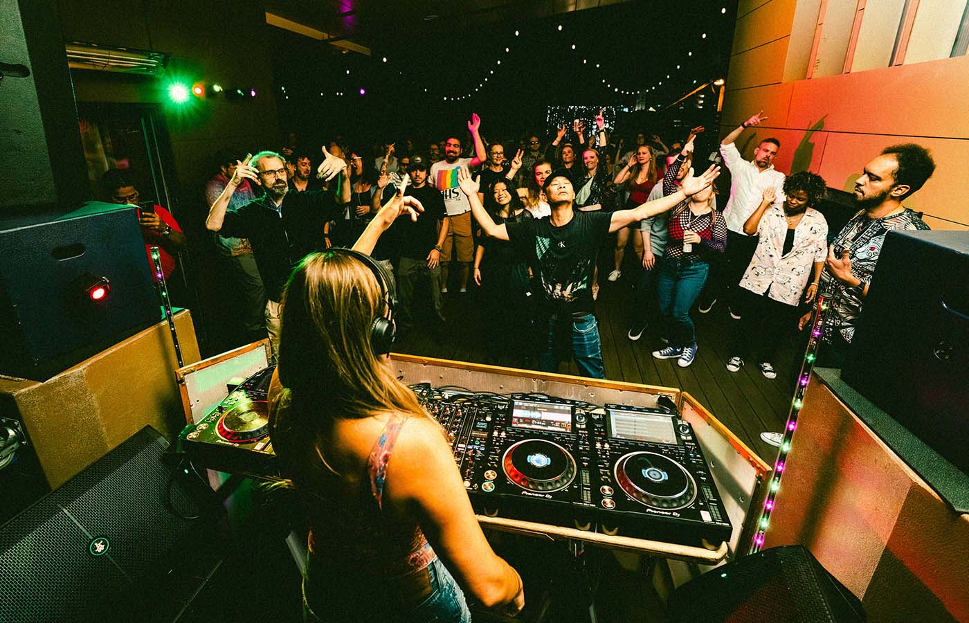 A DJ is performing in front of a lively crowd under colorful lights in a club-like setting. The audience is engaged and raising their hands, with a backdrop of string lights and vibrant decor.