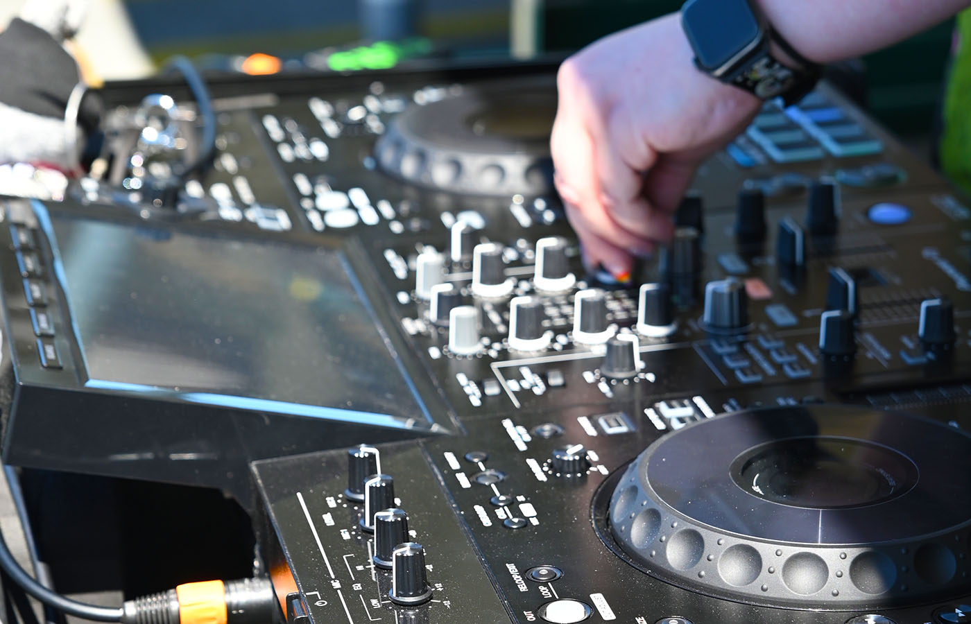 A person adjusting controls on a professional music mixing console, focusing on various knobs and sliders. Their wrist displays a smartwatch, and a tattoo is visible on their arm. The equipment is detailed and technical, suggesting a DJ setup.