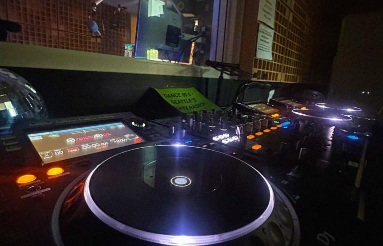 A dimly lit DJ booth featuring two illuminated turntables and a mixer with various knobs and buttons. A sign in the background mentions "Dance Hits - Seattle's Party Radio.