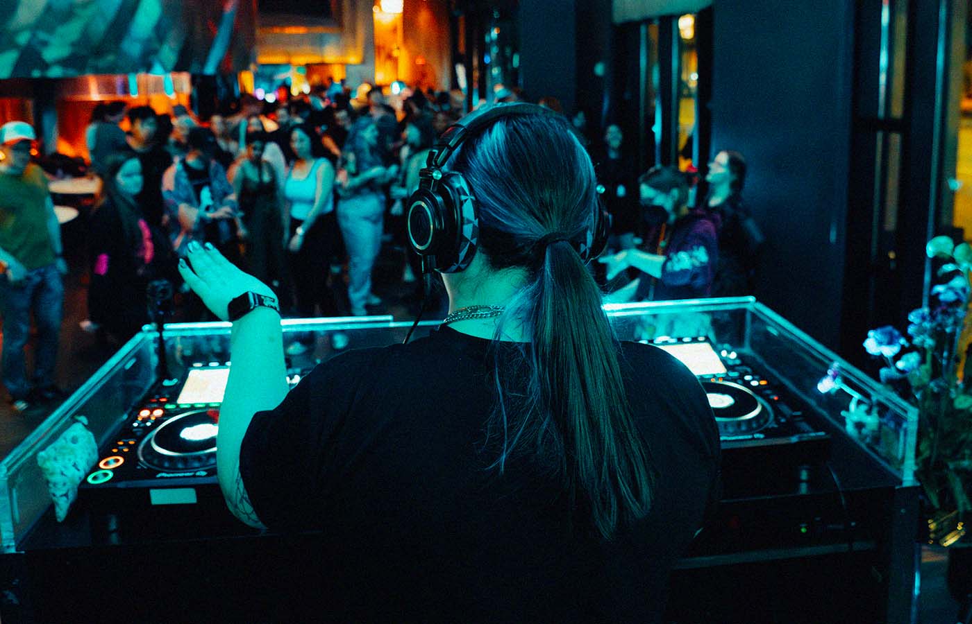 A DJ with headphones stands at a booth, facing a lively crowd in a dimly lit venue. The audience appears engaged, with some people taking photos or dancing. The setting is vibrant, with colorful lights creating an energetic atmosphere.