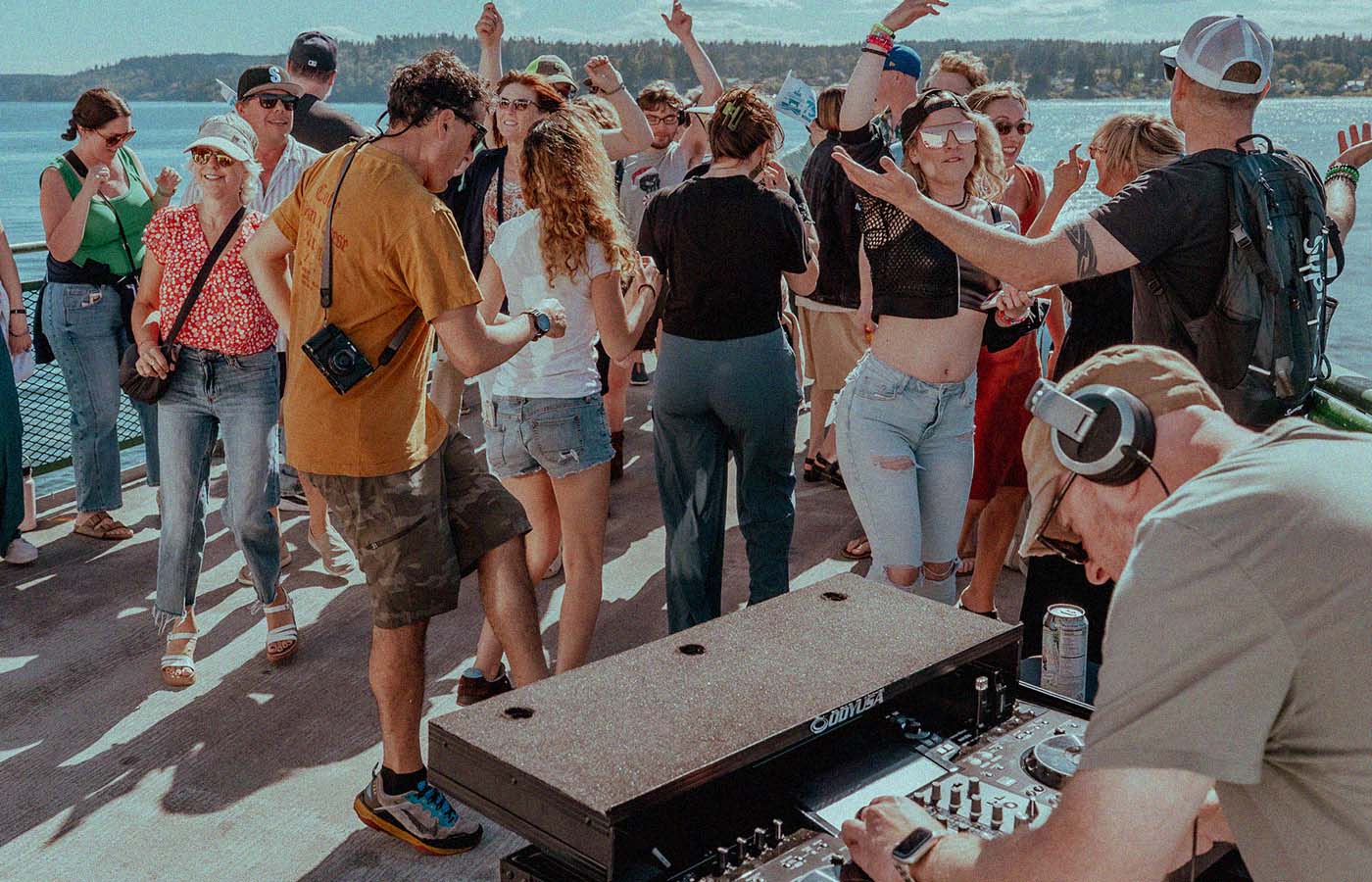 A group of people enjoying a lively outdoor gathering on a boat deck. A DJ is playing music from a mixer. The crowd is dancing and socializing under a sunny sky.