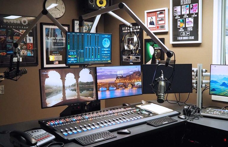 A modern radio studio with multiple computer monitors displaying various screens. There are two large microphones and a sound mixing console. The walls are decorated with awards, clocks, and framed posters.