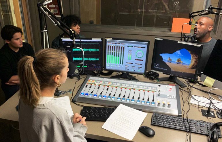 Four individuals are in a radio studio with sound equipment. One person is seated behind a desk with multiple screens and a mixing console, speaking into a microphone. Three others are seated nearby, listening attentively. The studio environment suggests a recording or broadcasting session.