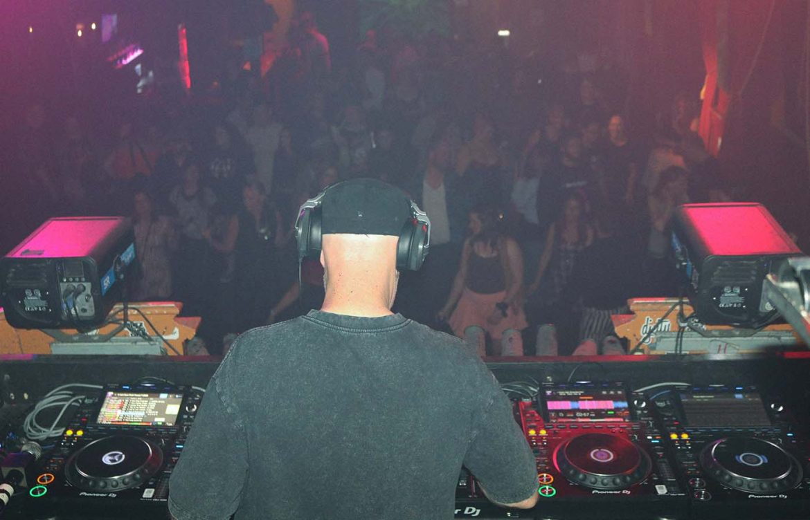 A person wearing a black cap and headphones is performing as a DJ in a dimly lit club. They are standing behind a DJ console with various controls and equipment. In the background, a large crowd is visible, dancing and enjoying the music. The lighting creates a vibrant atmosphere, and a "JM" logo is visible on a screen.