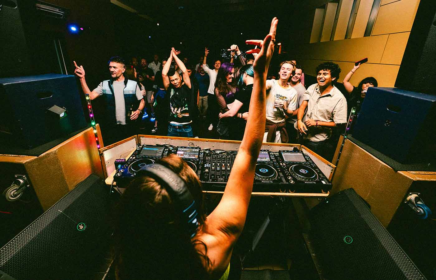 A DJ wearing headphones performs at a nightclub, with a lively crowd dancing and cheering in front of the booth. Multicolored lights illuminate the scene, and various DJ equipment is visible on the table.